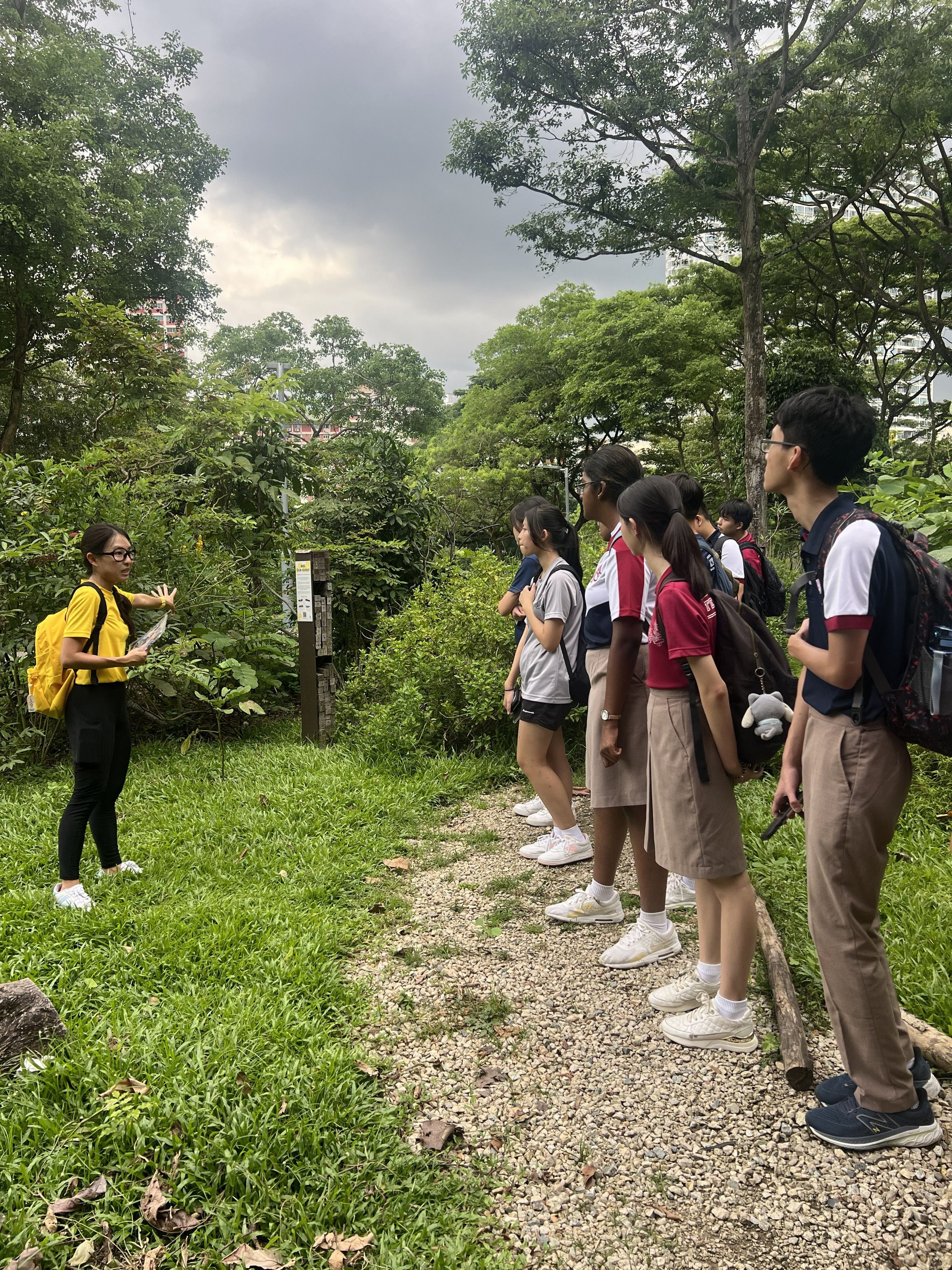 A guide in a yellow top addresses a group of students with backpacks along a gravel path surrounded by lush greenery at Bishan-Ang Mo Kio Park, under an overcast sky.
